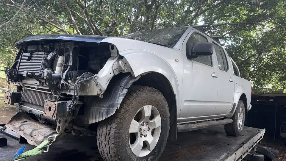 Rusted old car in a wrecking yard with trees and clear sky in the background.